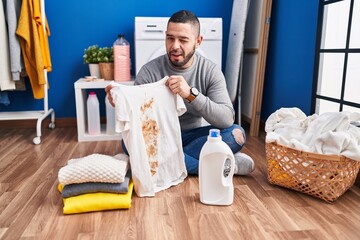 Hispanic man holding dirty t shirt with stain winking looking at the camera with sexy expression,...