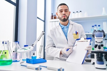 Hispanic man working at scientist laboratory holding blank clipboard looking at the camera blowing a kiss being lovely and sexy. love expression.