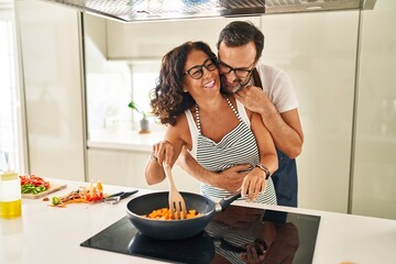 Middle age hispanic couple smiling confident and hugging each other cooking at kitchen