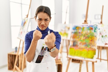 Young brunette woman at art studio ready to fight with fist defense gesture, angry and upset face, afraid of problem