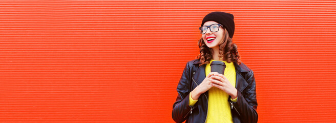 Portrait of stylish smiling young woman with cup of coffee wearing black rock style leather jacket, hat, eyeglasses on red background