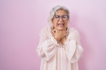 Middle age woman with grey hair standing over pink background shouting and suffocate because painful strangle. health problem. asphyxiate and suicide concept. - obrazy, fototapety, plakaty