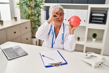 Middle age woman with grey hair wearing doctor uniform holding balloon smiling happy doing ok sign with hand on eye looking through fingers