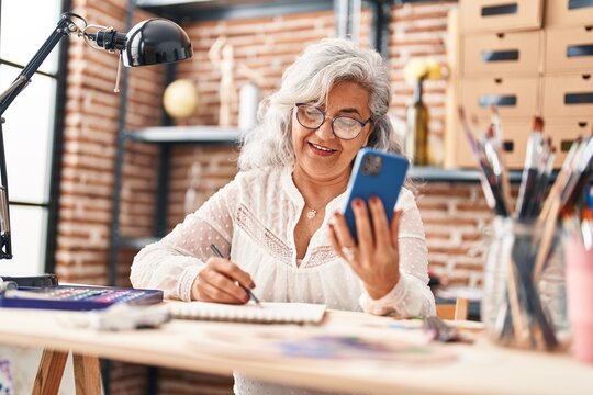 Middle age woman artist drawing on notebook using smartphone at art studio