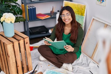 Young hispanic woman artist reading book sitting on floor at art studio