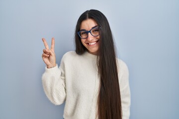 Young hispanic woman wearing casual sweater over blue background smiling with happy face winking at...