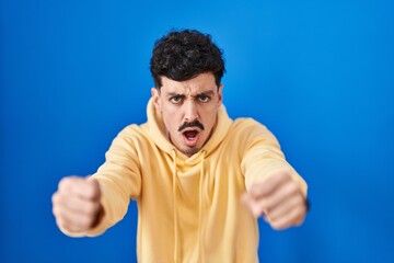 Hispanic man standing over blue background angry and mad raising fists frustrated and furious while shouting with anger. rage and aggressive concept.