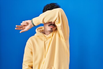 Hispanic man standing over blue background covering eyes with arm, looking serious and sad. sightless, hiding and rejection concept