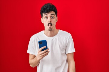 Hispanic man using smartphone over red background making fish face with lips, crazy and comical gesture. funny expression.