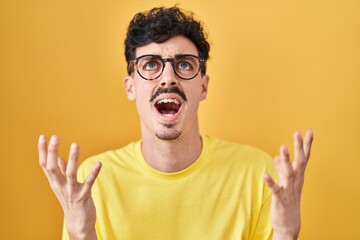 Hispanic man wearing glasses standing over yellow background crazy and mad shouting and yelling...