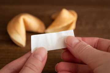 Person holds in hands blank paper slip from fortune cookie against two cookies laying on table surface background, mockup for your good luck wish