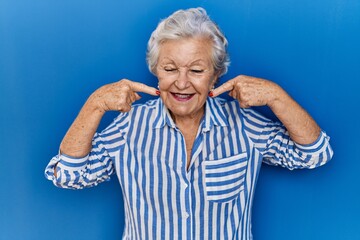 Senior woman with grey hair standing over blue background smiling cheerful showing and pointing with fingers teeth and mouth. dental health concept.
