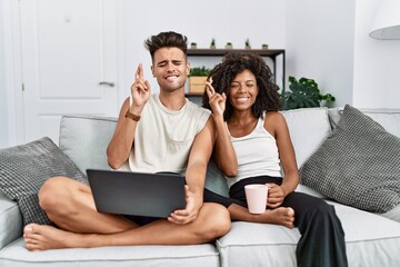 Young interracial couple using laptop at home sitting on the sofa gesturing finger crossed smiling...