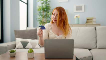 Young redhead woman using laptop and credit card sitting on sofa at home