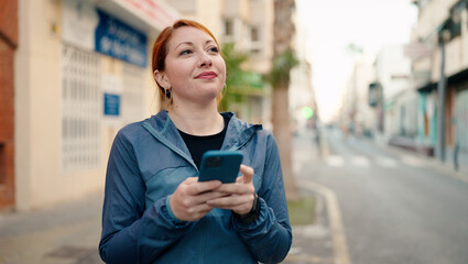 Young redhead woman wearing sportswear using smartphone at street