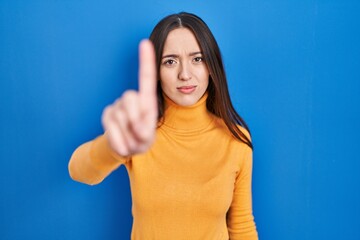 Fototapeta premium Young brunette woman standing over blue background pointing with finger up and angry expression, showing no gesture