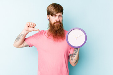 Young caucasian man holding a clock isolated on blue background feels proud and self confident,...