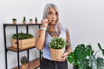 Middle age grey-haired woman holding green plant pot at home mouth and lips shut as zip with fingers. secret and silent, taboo talking