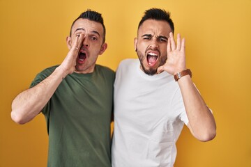 Homosexual couple standing over yellow background shouting and screaming loud to side with hand on mouth. communication concept.
