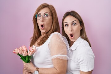 Hispanic mother and daughter holding pink tulips bouquet scared and amazed with open mouth for surprise, disbelief face