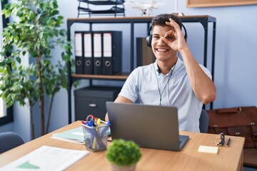 Young hispanic man working at the office wearing headphones doing ok gesture with hand smiling, eye looking through fingers with happy face.