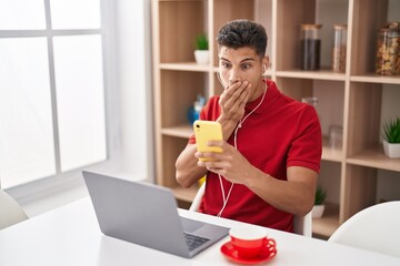 Young hispanic man using laptop and doing video call with smartphone covering mouth with hand,...