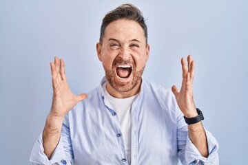 Middle age caucasian man standing over blue background crazy and mad shouting and yelling with...