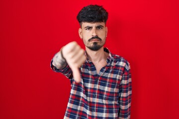 Fototapeta premium Young hispanic man with beard standing over red background looking unhappy and angry showing rejection and negative with thumbs down gesture. bad expression.
