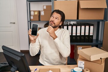 Handsome middle age man working at small business ecommerce holding smartphone serious face...