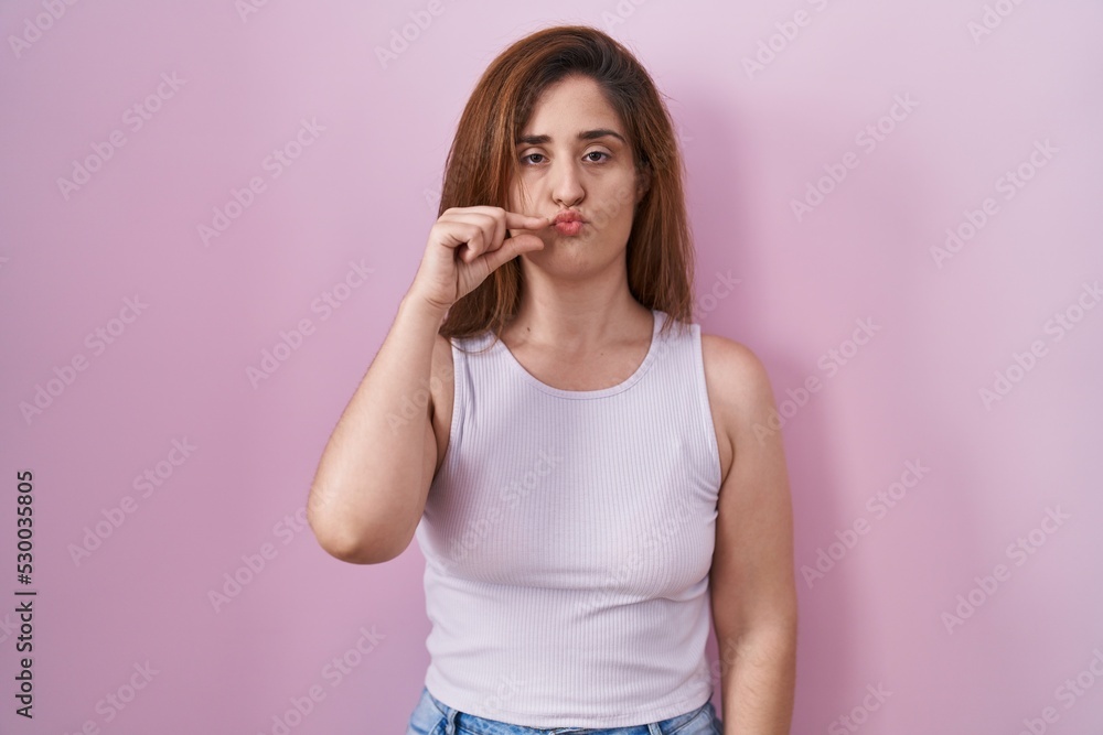 Poster brunette woman standing over pink background mouth and lips shut as zip with fingers. secret and sil