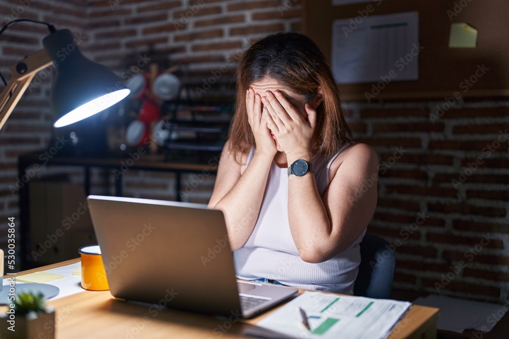 Wall mural Brunette woman working at the office at night with sad expression covering face with hands while crying. depression concept.