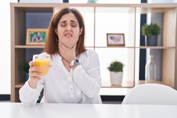 Brunette woman drinking glass of orange juice touching painful neck, sore throat for flu, clod and infection