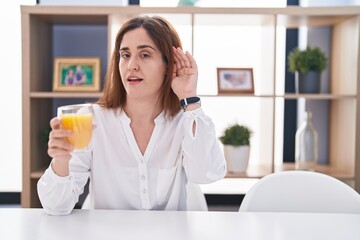 Brunette woman drinking glass of orange juice smiling with hand over ear listening an hearing to rumor or gossip. deafness concept.