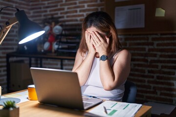 Brunette woman working at the office at night with sad expression covering face with hands while...