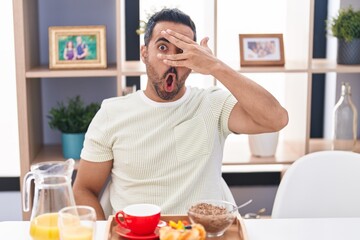 Hispanic man with beard eating breakfast peeking in shock covering face and eyes with hand, looking through fingers afraid