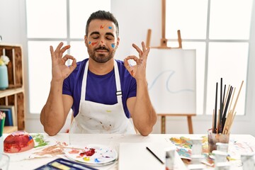 Young hispanic man with beard at art studio with painted face relax and smiling with eyes closed doing meditation gesture with fingers. yoga concept.