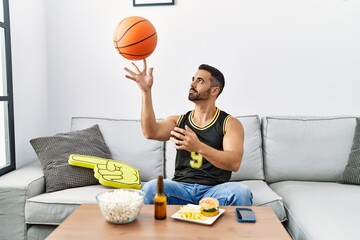 Young hispanic man supporting basketball game playing with ball at home
