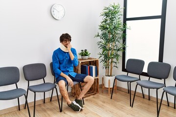 Young hispanic man sitting at doctor waiting room with neck injury looking stressed and nervous with hands on mouth biting nails. anxiety problem.