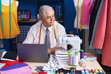 Middle age grey-haired man tailor using sewing machine and laptop at tailor shop