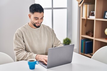 Fototapeta premium Young arab man using laptop and drinking coffee sitting on table at home