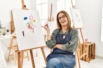 Middle age artist woman at art studio with a big smile on face, pointing with hand and finger to...