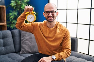 Young bald man with beard holding alarm clock looking positive and happy standing and smiling with...