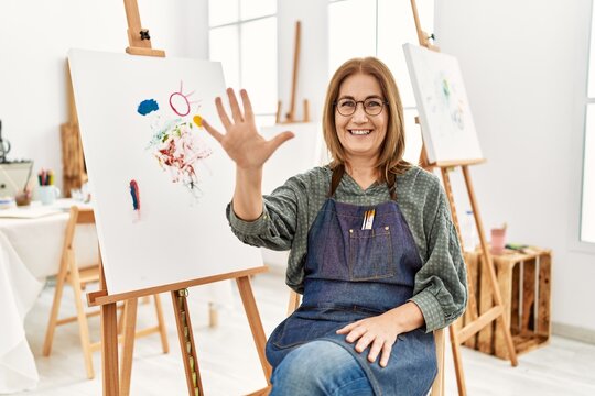 Middle age artist woman at art studio showing and pointing up with fingers number five while smiling confident and happy.