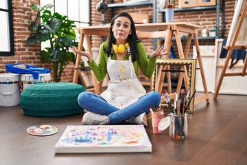 Hispanic woman sitting at art studio painting on canvas clueless and confused expression with arms and hands raised. doubt concept.