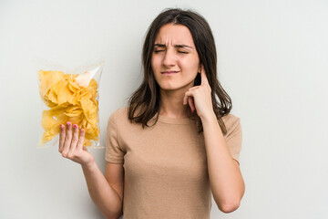 Young caucasian woman holding crisps isolated on yellow background covering ears with hands.