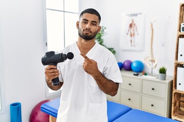 Young indian physiotherapist holding therapy massage gun at wellness center pointing with hand finger to the side showing advertisement, serious and calm face