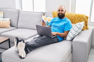 Young bald man using laptop and credit card sitting on sofa at home