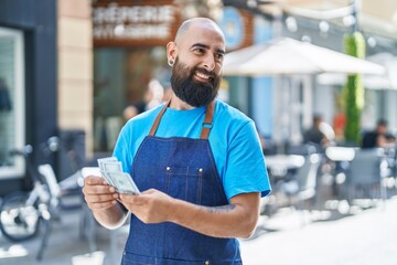 Young bald man waiter smiling confident counting dollars at coffee shop terrace