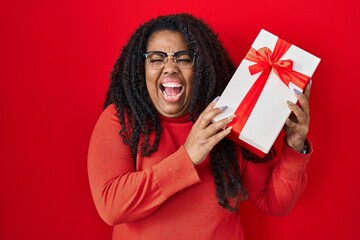Plus size hispanic woman holding gift celebrating crazy and amazed for success with open eyes screaming excited.