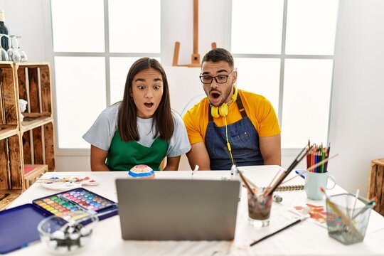 Young hispanic couple using laptop sitting on the table at art studio scared and amazed with open mouth for surprise, disbelief face